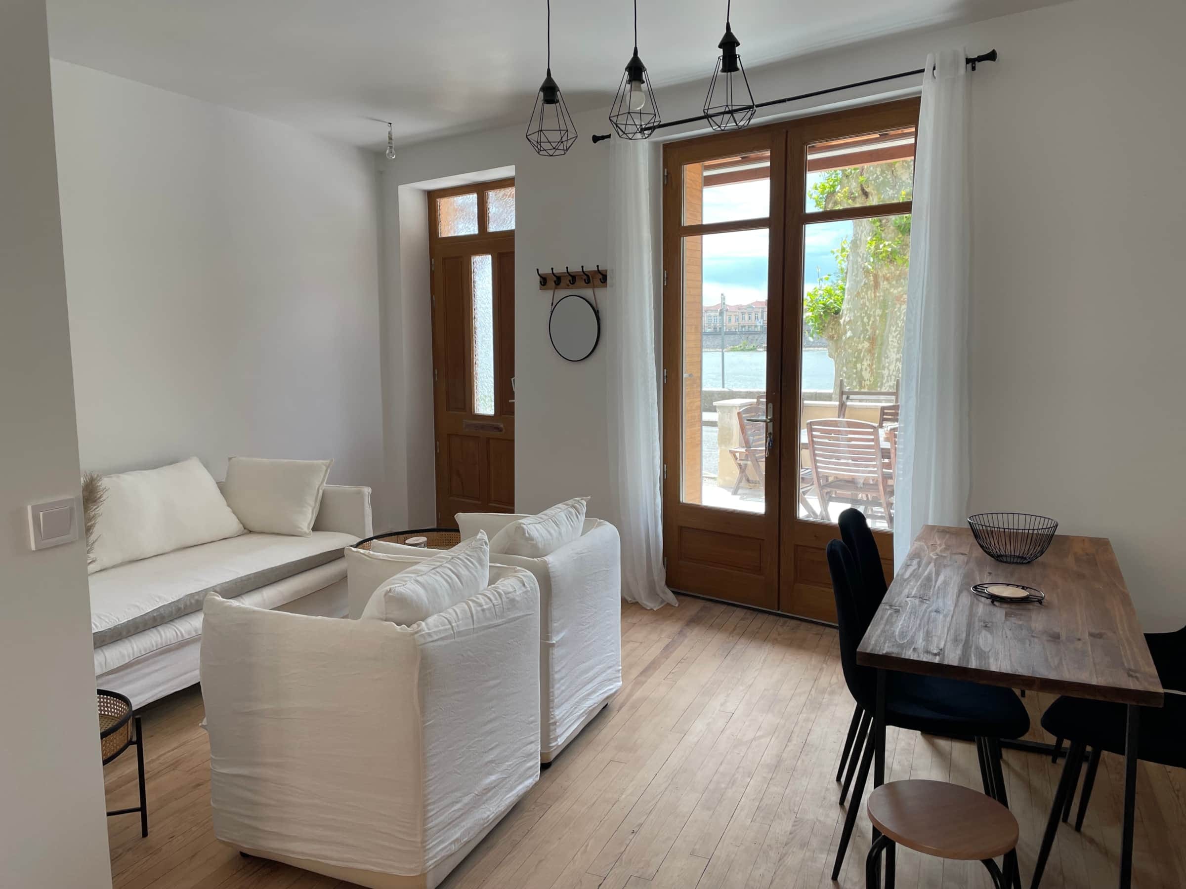 Séjour de la maison de vacances avec banquette, fauteuils blancs, table à manger et porte-fenêtre sur le quai